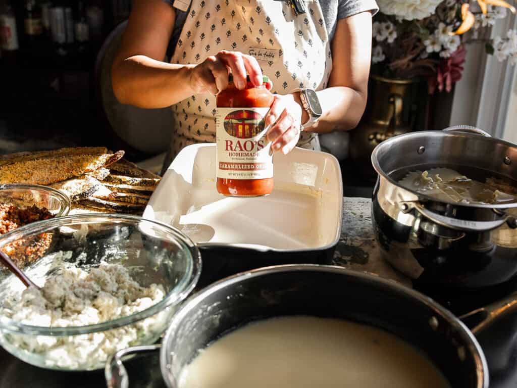 Person preparing a dish by spreading Raos tomato sauce in a rectangular baking dish. Various ingredients like cheese and pasta are arranged around, and theres a pot of white sauce nearby. The setting suggests a cozy kitchen environment.